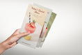 Load image into Gallery viewer, Hand holding some Flyers with a strawberry dessert illustration on a light gray background
